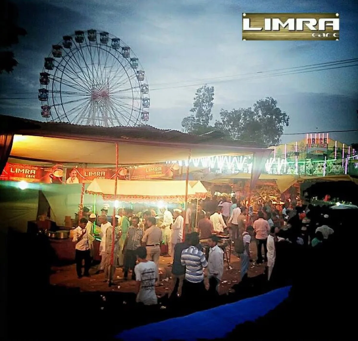 A crowd at the Limra Café stall at dusk, with Limra banners overhead and a Ferris wheel in the background